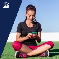 Eine sportlich bekleidete Frau sitzt auf einem Rasen und bedient ein Smartphone